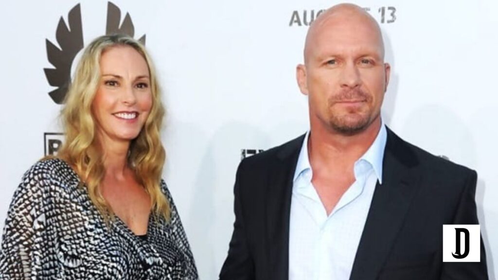 A smiling blonde woman in a patterned dress poses beside a bald man in a black suit at a formal event with a white backdrop